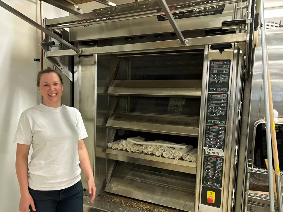 Maria Holbrow stands proudly in front of a large commercial oven in her bakery, ready to bake many loaves of bread.