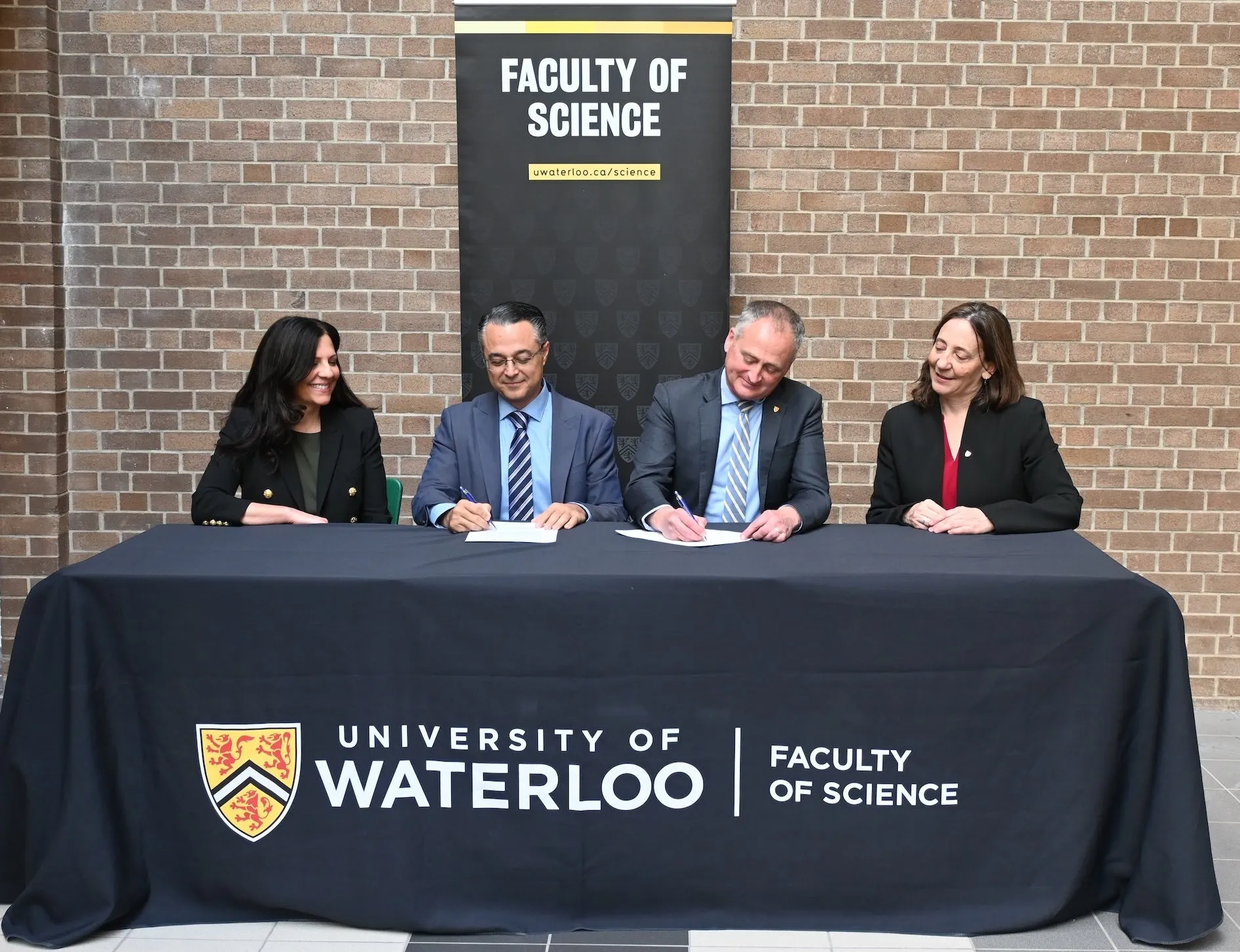 Four people, two men and two women, seated around a table signing documents during a university partnership agreement