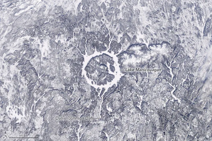 Satellite image in natural color shows a vast, icy white ring lake surrounded by gray and white snowy terrain.