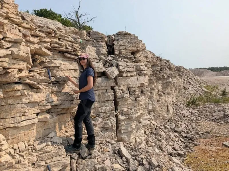 Dr. Melina Jobbins stands in a quarry near Lake Manitoba, pointing to a rock layer.